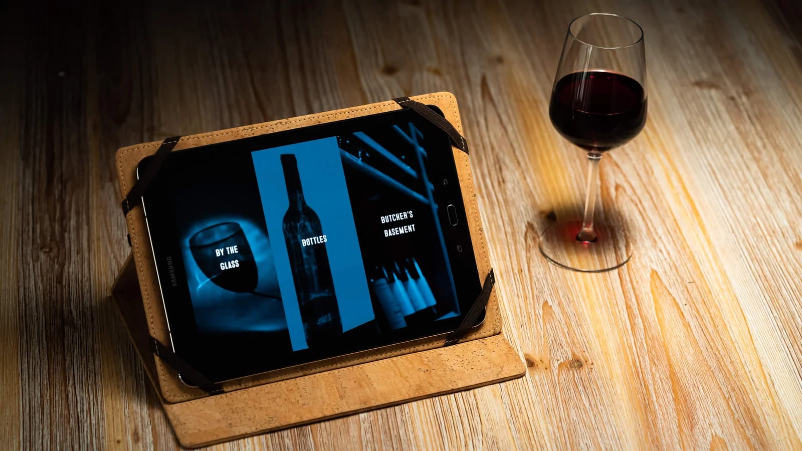 De wijnkaart van Bar Paloma op een tablet in een kurken hoes, op een houten tafel naast een glas rode wijn.